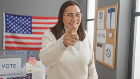 Mujer hispana señalando dentro de un colegio electoral de EE.UU. con bandera americana, sonriente y confiada.