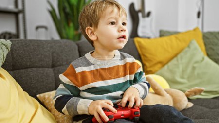 Photo for A focused blond boy playing with a game controller in a cozy living room, surrounded by colorful pillows and a plush toy. - Royalty Free Image