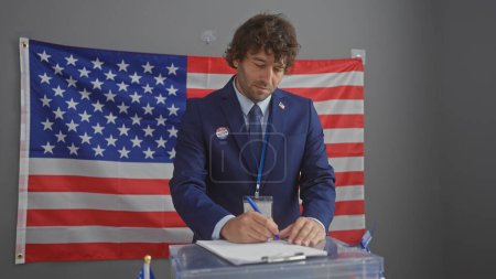 A young man with a beard in a suit writes on a paper in an indoor setting with an american flag backdrop, depicting a voting scene.