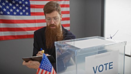 Hombre barbudo con gafas tomando notas al lado de una urna con banderas americanas en una estación de votación.