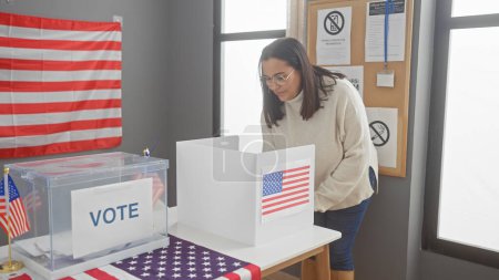 Foto de Una mujer hispana de mediana edad vota adentro en un centro electoral de EE.UU. con una bandera americana visible. - Imagen libre de derechos