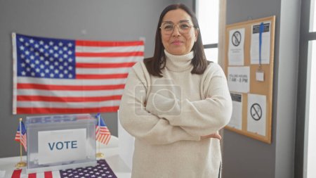 Middle-aged hispanic woman with arms crossed in an indoor us electoral center with american flag