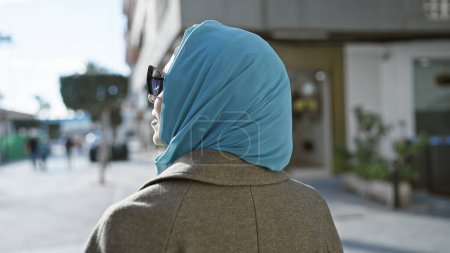 Foto de Una mujer madura en un hijab y gafas de sol camina por una calle urbana soleada, exudando una sensación de confianza y elegancia. - Imagen libre de derechos