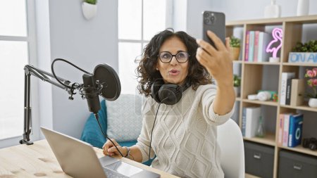 Frau mittleren Alters im Livestream in einem modernen Radiostudio mit Mikrofon und Laptop.
