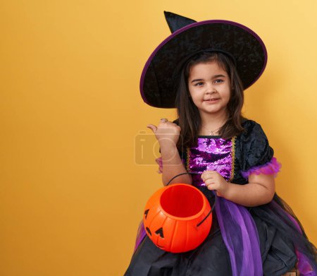 Photo for Adorable hispanic girl in witch costume, joyously flashing a thumbs-up, pointing to side with a pumpkin basket, and a cheerful, open-mouthed smile on yellow isolated background. - Royalty Free Image