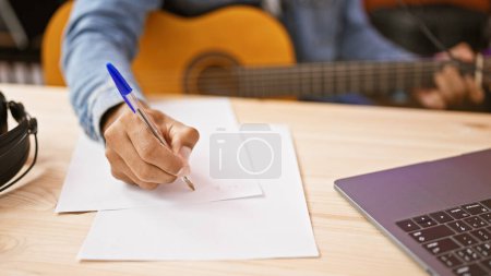 Téléchargez les photos : Homme hispanique écrivant des notes de studio de musique avec guitare et ordinateur portable - en image libre de droit