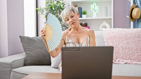 A mature woman sits on her sofa indoors, holding a fan, and using a laptop with stylish decor in the background.