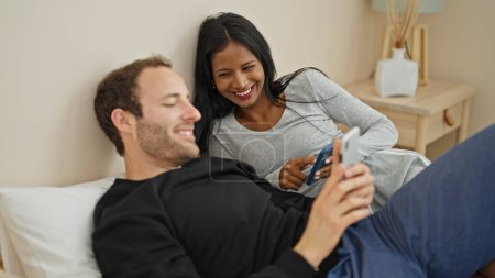 Téléchargez les photos : Beau couple couché sur le lit en utilisant des smartphones souriant à la chambre - en image libre de droit