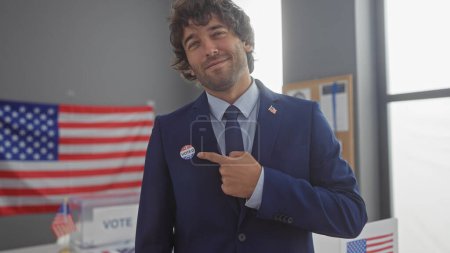 Handsome young hispanic man with beard wearing a suit at an american electoral college, pointing to 'i voted' sticker with a proud smile