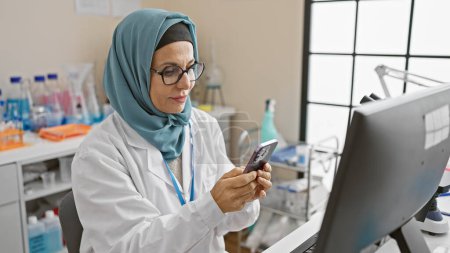 Foto de Científica de mediana edad en hijab examina datos sobre teléfonos inteligentes en laboratorio - Imagen libre de derechos