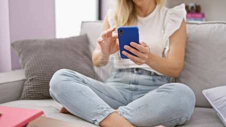 Photo for Young blonde woman using smartphone sitting on sofa at home - Royalty Free Image