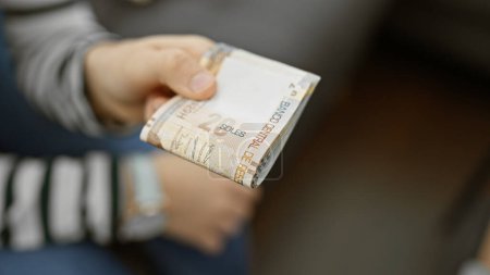 Foto de La mano de una mujer sosteniendo billetes de suela peruana con un fondo interior borroso que sugiere un ambiente hogareño. - Imagen libre de derechos