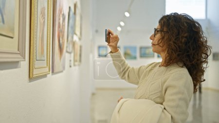 Foto de Mujer hispana madura tomando fotos en galería de arte con pinturas - Imagen libre de derechos