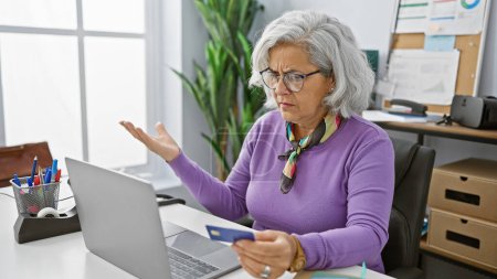 Una mujer de mediana edad perpleja con el pelo gris examina su tarjeta de crédito en una oficina moderna.