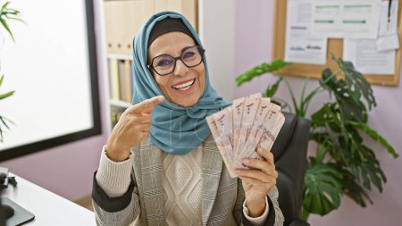 Smiling woman in hijab pointing at saudi riyals in a modern office.
