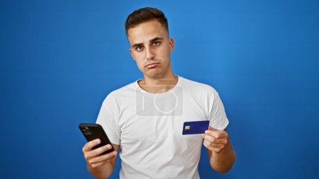 Worried young adult hispanic man holding smartphone and credit card against isolated blue background.