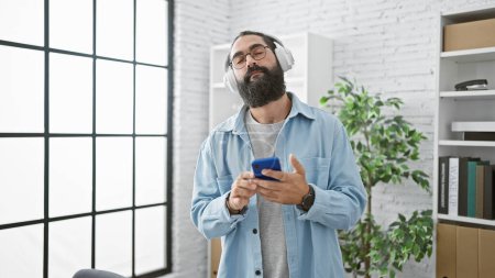 A relaxed hispanic man in a modern office enjoys music on his headphones while using a smartphone.