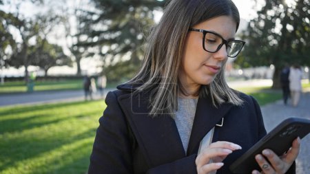 Foto de Mujer latina adulta usando gafas utiliza un teléfono inteligente en un parque urbano con vegetación borrosa y personas. - Imagen libre de derechos