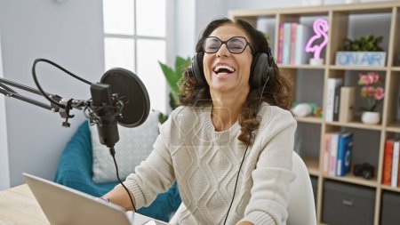 Mujer sonriente usando auriculares podcasting en un estudio en casa, con un micrófono y un ordenador portátil.