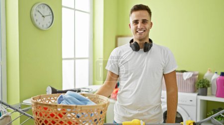 Téléchargez les photos : Beau homme avec écouteurs dans une buanderie lumineuse à la maison, souriant tout en faisant des travaux ménagers. - en image libre de droit