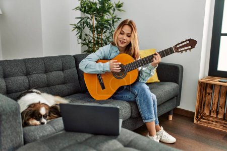 Téléchargez les photos : Jeune femme caucasienne ayant cours de guitare classique en ligne assis sur un canapé avec chien à la maison - en image libre de droit