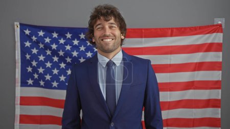 Lächelnder junger hispanischer Mann im Anzug vor amerikanischer Flaggenkulisse drinnen, der ein professionelles, multikulturelles Umfeld darstellt.