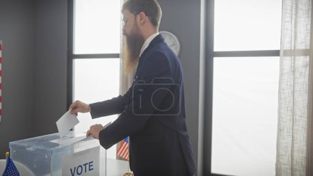 Photo for A bearded man in a suit casts a ballot into a transparent voting box in a room with american flags, exemplifying democracy. - Royalty Free Image