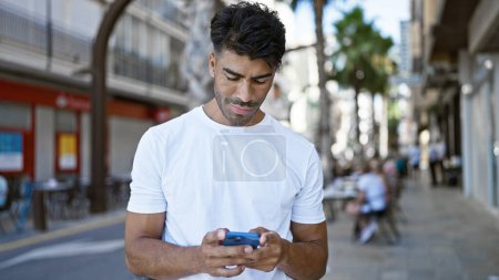 Schöner hispanischer Mann textet auf Smartphone Innenstadt, urbane Lifestyleszene mit verschwommenem Stadthintergrund.