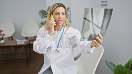 Foto de Mujer rubia joven médico buscando rayos X hablando en el teléfono inteligente en la sala de espera clínica - Imagen libre de derechos