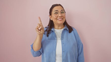 Téléchargez les photos : Une femme hispanique souriante d'âge moyen avec des lunettes pointées vers le haut, sur un fond rose isolé. - en image libre de droit