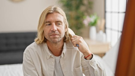 Téléchargez les photos : Beau homme toilettant de longs cheveux blonds avec un peigne dans une chambre lumineuse - en image libre de droit