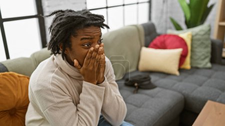 Foto de Sorprendida joven mujer negra con rastas sentada en un sofá en un acogedor salón interior. - Imagen libre de derechos