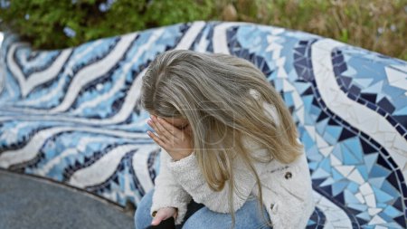 Photo for Young woman sitting outdoors on a mosaic bench, covering her face with her hand in a park setting - Royalty Free Image