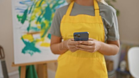 Foto de Una mujer en un estudio se centra en su teléfono con una pintura en el fondo, que encarna la creatividad y la conectividad moderna. - Imagen libre de derechos