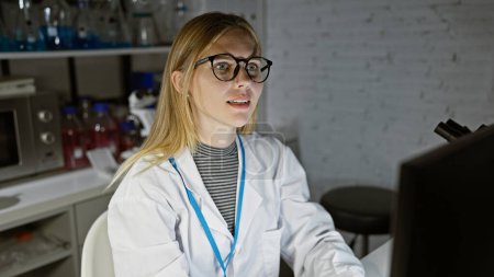 Foto de Una mujer rubia en gafas trabaja con atención en un entorno de laboratorio, rodeada de equipo científico. - Imagen libre de derechos