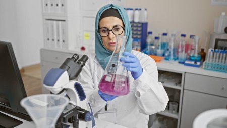 Photo for A focused woman scientist examines a flask in a laboratory, surrounded by scientific equipment and a microscope. - Royalty Free Image