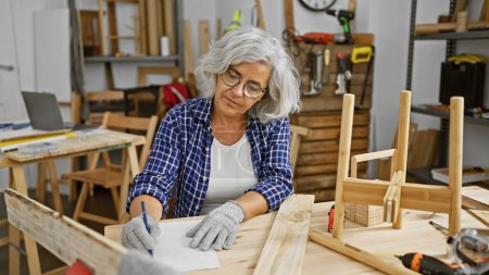 Photo for Mature woman in glasses plans woodworking project in a workshop - Royalty Free Image