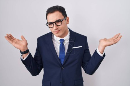 Photo for Young hispanic man wearing suit and tie clueless and confused expression with arms and hands raised. doubt concept. - Royalty Free Image