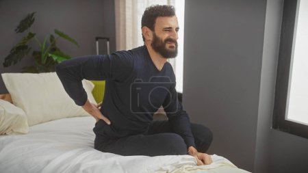 Foto de Un hombre barbudo con atuendo casual hace muecas con dolor de espalda mientras está sentado en una cama blanca en el interior de un dormitorio moderno. - Imagen libre de derechos
