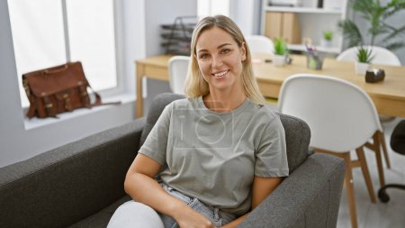 Photo for A young, smiling, caucasian woman sits casually in a modern office, exuding confidence and approachability. - Royalty Free Image