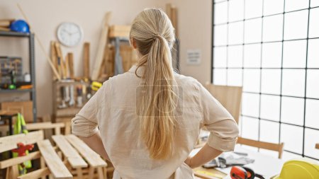 Téléchargez les photos : Vue arrière d'une jeune femme blonde dans un atelier de menuiserie artisanale, entourée d'équipements de menuiserie. - en image libre de droit