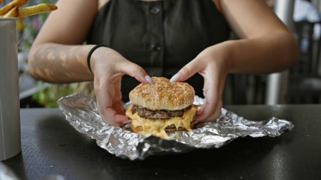 Téléchargez les photos : Gros plan des mains d'une femme tenant un cheeseburger en plein air, offrant une expérience culinaire décontractée. - en image libre de droit
