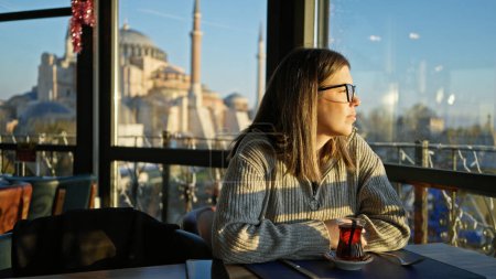 A contemplative woman enjoys tea in a restaurant with a view of hagia sophia in istanbul at sunset. Stickers 710166498