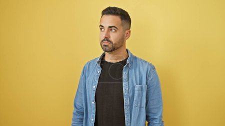 Photo for Handsome hispanic man with beard in casual denim jacket against yellow background, looking aside thoughtfully. - Royalty Free Image