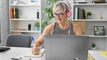 Téléchargez les photos : Une femme mûre souriante avec des écouteurs utilisant un ordinateur portable et prenant des notes dans un environnement de bureau. - en image libre de droit