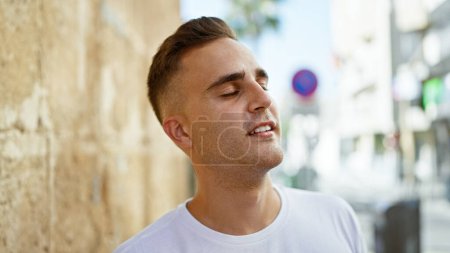 Téléchargez les photos : Beau jeune homme hispanique en chemise blanche décontractée profitant d'une journée ensoleillée sur une rue animée de la ville - en image libre de droit
