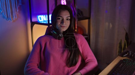Foto de Una joven hispana en un suéter rosa con auriculares se sienta en un escritorio en una sala de juegos oscura con luces de neón. - Imagen libre de derechos