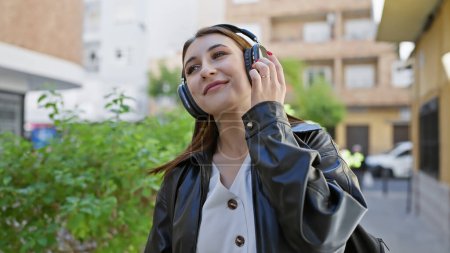 Téléchargez les photos : Une jeune femme brune aime la musique sur son casque tout en se promenant dans une rue urbaine, incarnant un style décontracté et de détente. - en image libre de droit