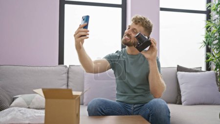 Junger, gutaussehender Mann mit Bart macht ein Selfie drinnen, während er in einem modernen Wohnzimmer eine Schachtel auspackt.