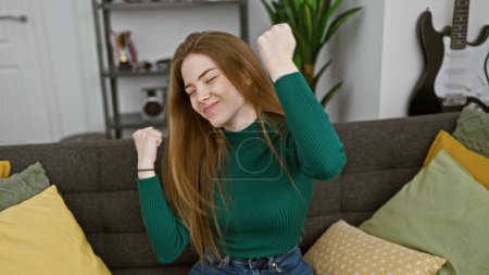 Foto de Una joven con el pelo rubio celebra exuberantemente en un acogedor ambiente de sala de estar. - Imagen libre de derechos
